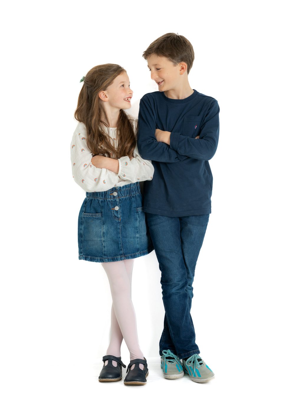 A young girl and boy stand together against a white background, facing and smiling at each other with arms folded. The girl wears a white blouse, denim skirt, white tights, and black shoes. The boy wears a navy long-sleeve top and blue jeans with light trainers.