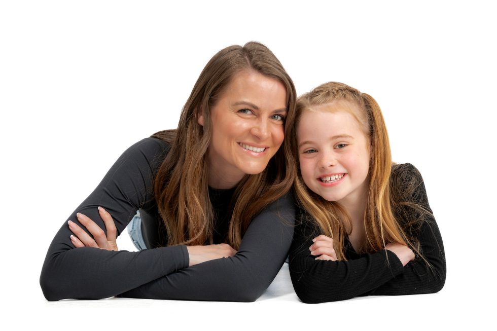 A woman and a young girl with long hair, both in black tops, lie side by side on a white background. They are smiling warmly at the camera, with their arms folded and heads close together, showing a close, affectionate bond.