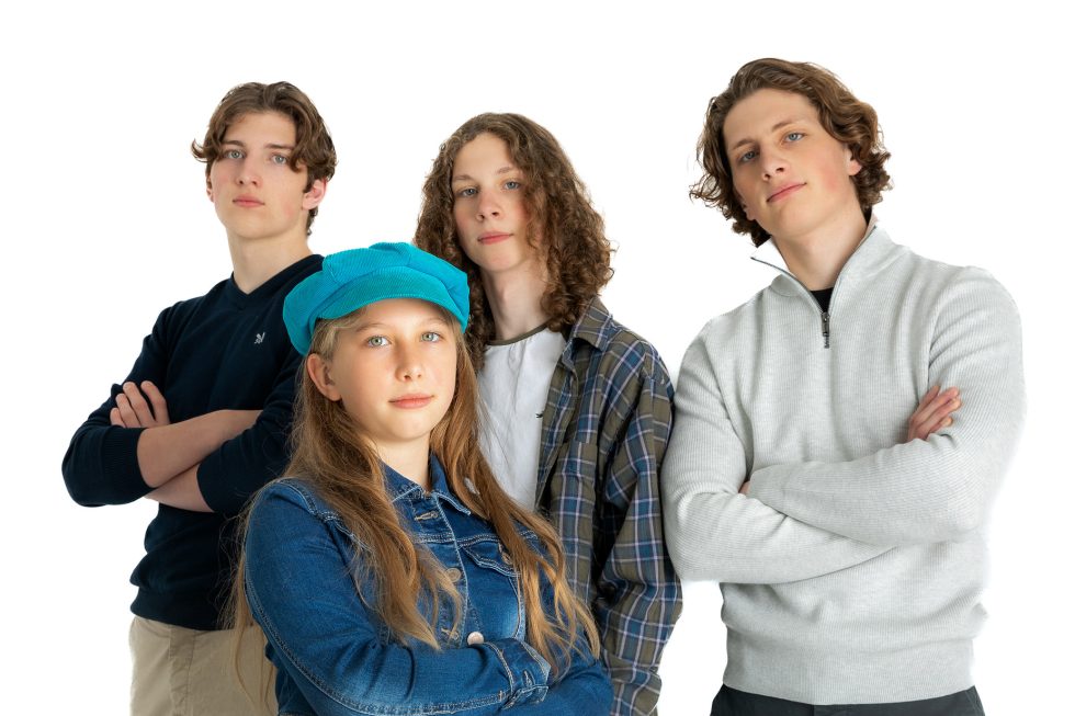 Four teenagers stand confidently against a white background, arms folded. The girl at the front wears a blue cap and denim jacket; behind her are three boys with wavy hair, wearing casual jumpers and shirts, all looking forward with relaxed expressions.