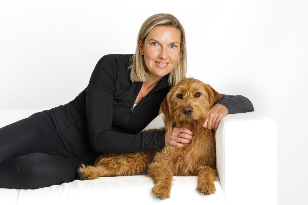 A smiling woman with blonde hair, wearing a black long-sleeved top and trousers, sits on a white sofa with her arm around a brown, shaggy dog. The woman and dog look directly at the camera against a plain white background.
