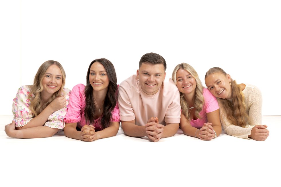 Five young adults lie side by side on the floor, elbows down and hands clasped, smiling at the camera against a white background. Four women wear pastel tops, and a man in the centre wears a pale pink shirt.