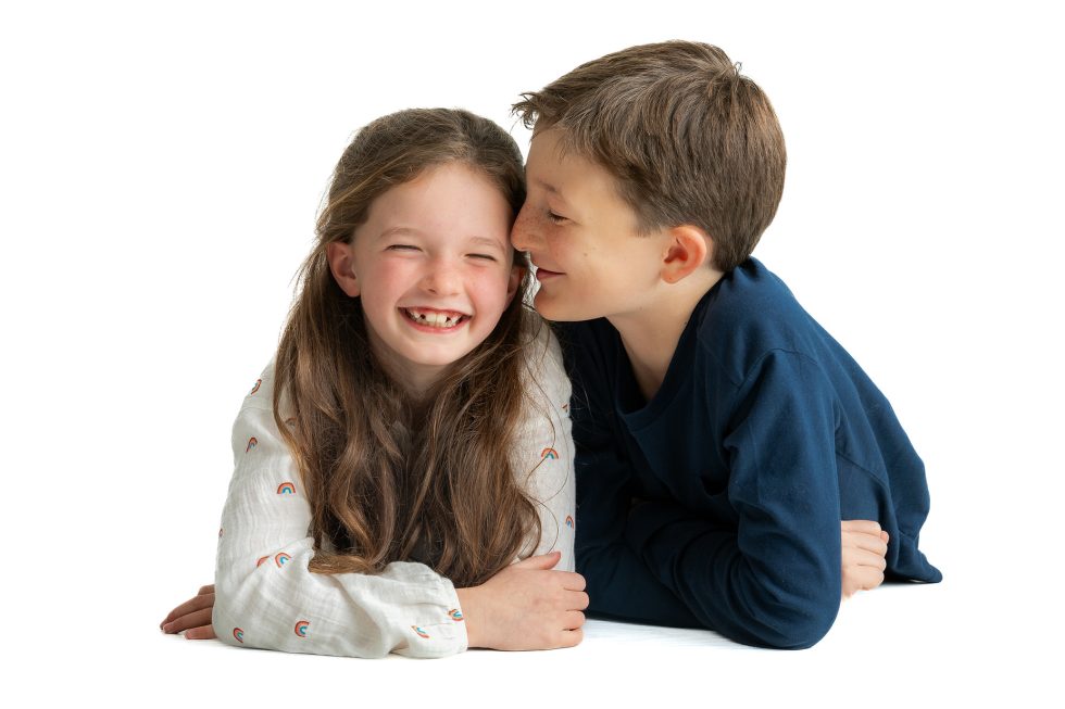 Two smiling children lie on the floor against a white background. The girl with long brown hair and a white shirt laughs, eyes closed, whilst the boy with short brown hair in a blue shirt leans in close, smiling affectionately at her.