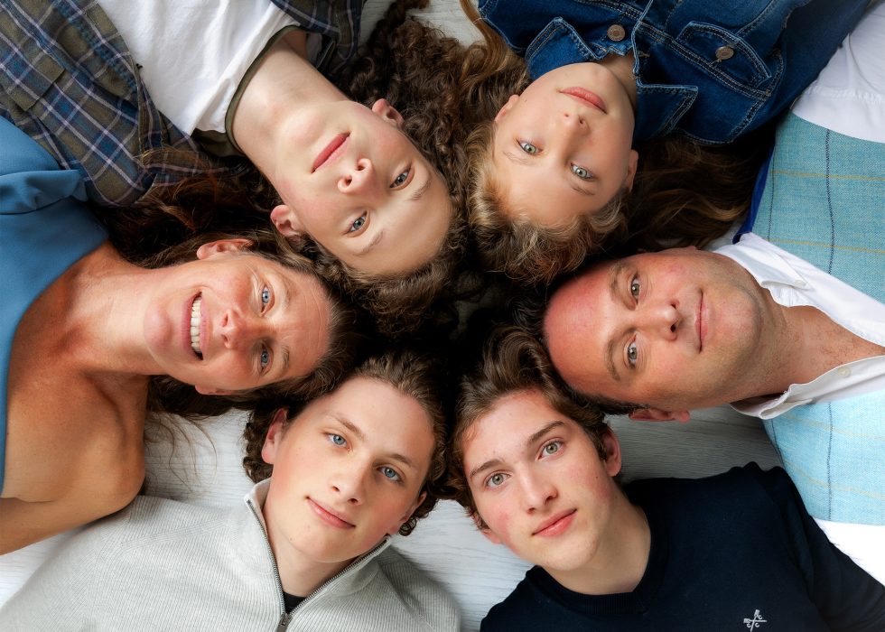 Six people, four teenagers and two adults, lie on the floor in a circle with their heads close together, looking up at the camera. They all have light skin and blue or green eyes, and they are smiling or have neutral expressions.