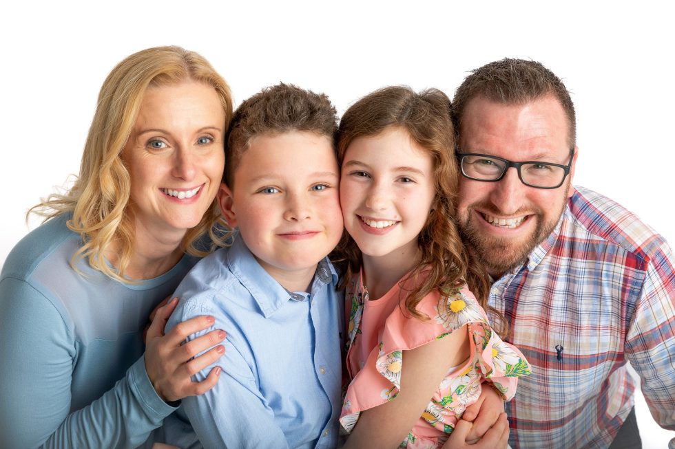 A smiling family of four pose closely together against a white background. The mother has blonde hair and wears a light blue top, the father has glasses and a beard and wears a checked shirt, and their two children, a boy in a blue shirt and a girl in a floral dress, stand between them.