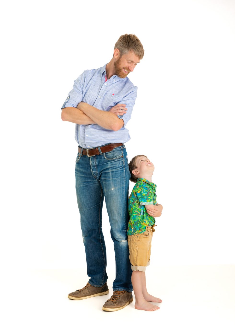 A bearded man in a blue shirt and jeans stands smiling with arms folded, looking down at a young barefoot boy in a green shirt and tan shorts. The boy looks up at the man, smiling. Both are standing against a plain white background.