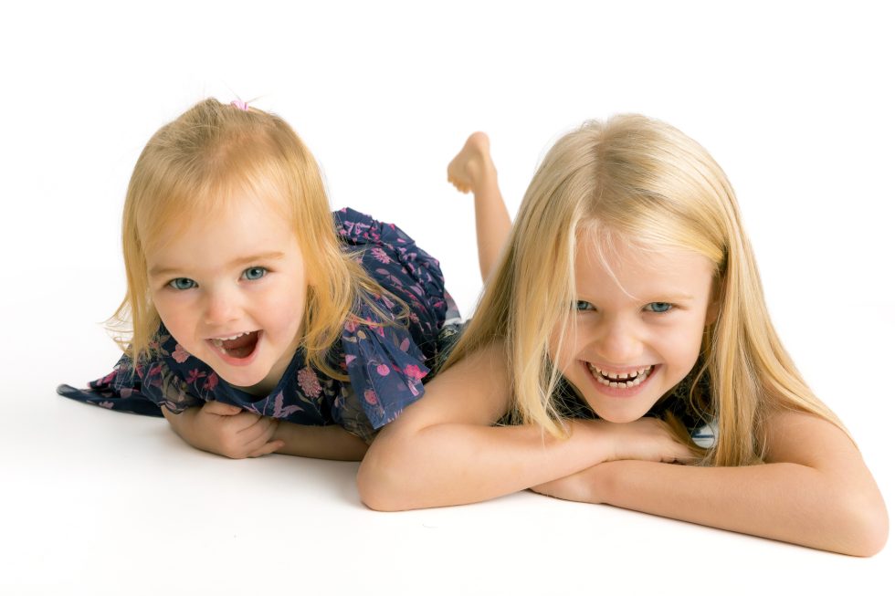 Two young girls with blonde hair are lying on the floor, facing the camera and smiling brightly. The girl on the left wears a blue, patterned dress, while the girl on the right is in a sleeveless top. They are against a white background.