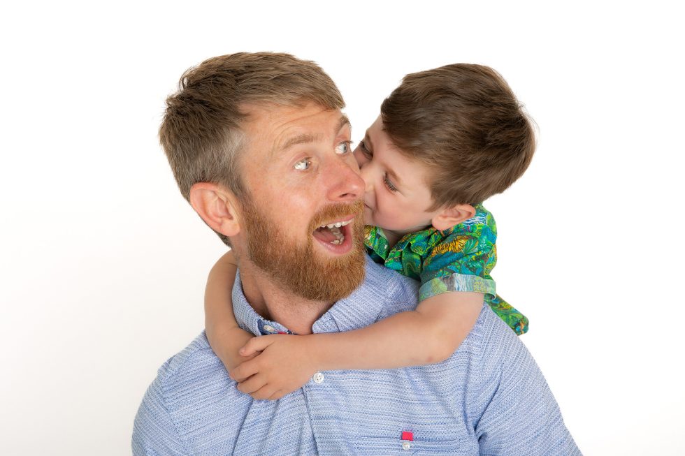 A bearded man in a light blue shirt smiles with surprise as a young boy in a colourful shirt hugs him from behind and playfully kisses his cheek. Both are against a plain white background, conveying warmth and affection.