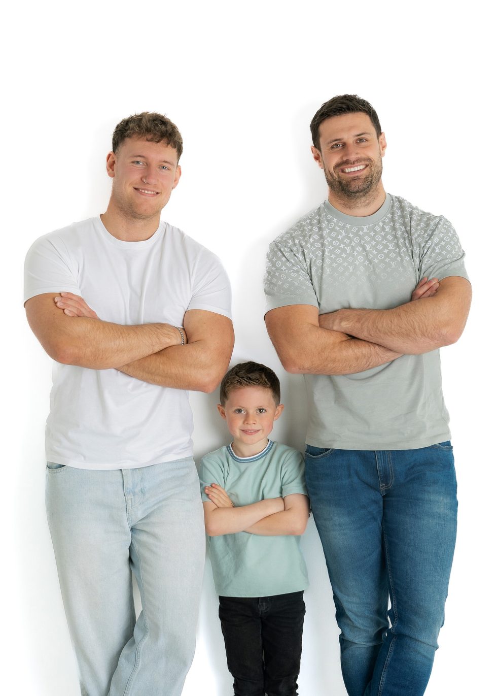 Three males stand against a white background. Two adults with muscular builds, wearing light t-shirts and jeans, stand on either side of a young boy in a light green t-shirt. All three have their arms folded and are smiling at the camera.