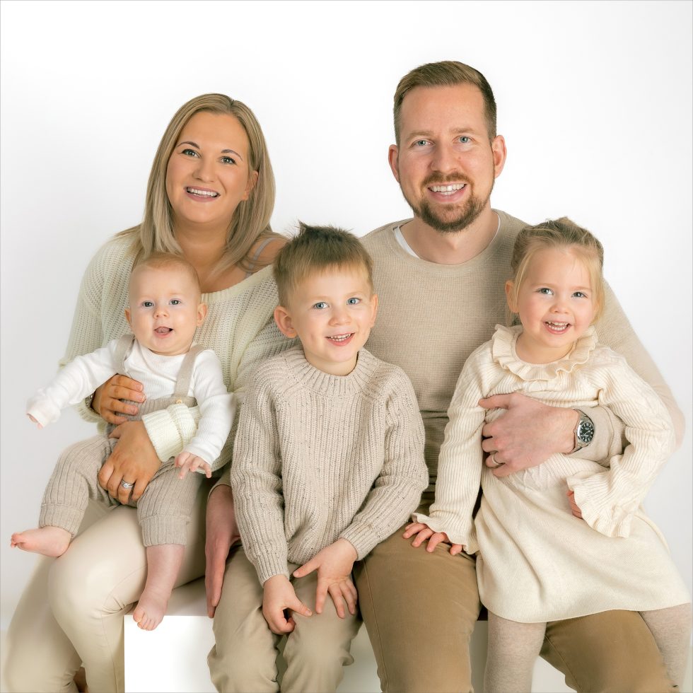 A smiling family of five poses in a studio against a white background. The mother holds a baby in white clothes, the father sits beside her, and two young children, a boy in a beige jumper and a girl in a cream dress, sit in front. All wear neutral, co-ordinated outfits.