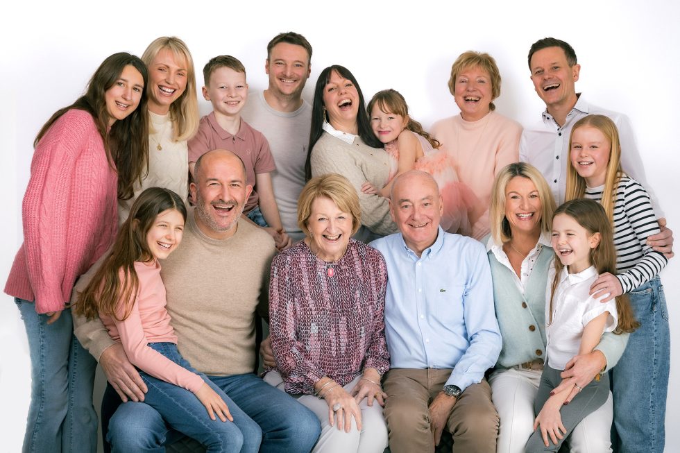 A multigenerational family of fifteen, including children, teenagers, and adults, pose closely together, smiling and laughing against a plain white background. They wear casual pastel and neutral clothing. Some are seated while others stand behind them.