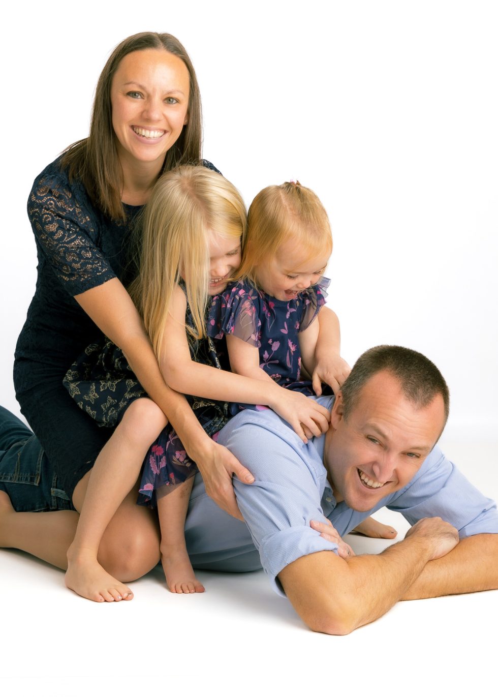 A happy family of four poses playfully on the floor. The father lies on his stomach, smiling, while two young daughters in matching dresses climb on his back. The mother, in a dark dress, kneels beside them, smiling at the camera against a white background.