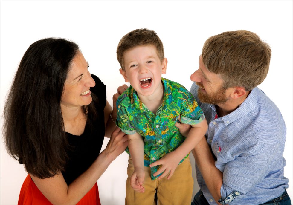 A smiling woman and man hold a laughing young boy between them. The boy wears a green patterned shirt and tan shorts; the adults are casually dressed. All three look happy against a plain white background, enjoying a playful moment together.