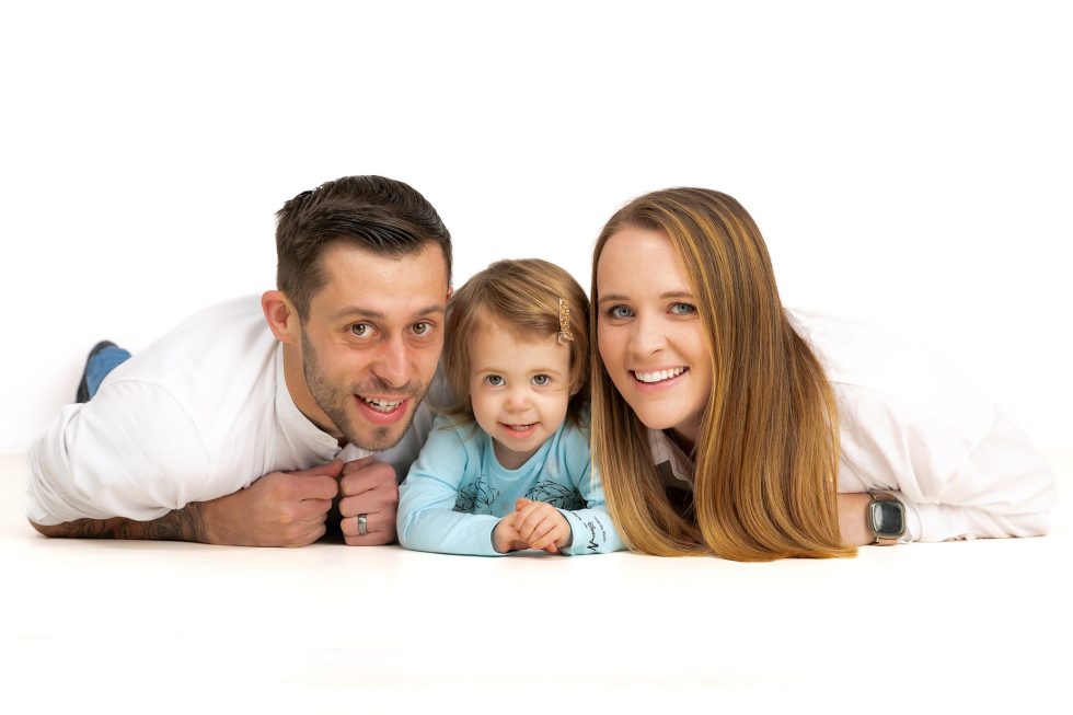 A man, a young girl, and a woman lie side by side on the floor, smiling at the camera in a brightly lit, white studio. The man and woman wear white tops, whilst the little girl wears light blue. All appear happy and relaxed.