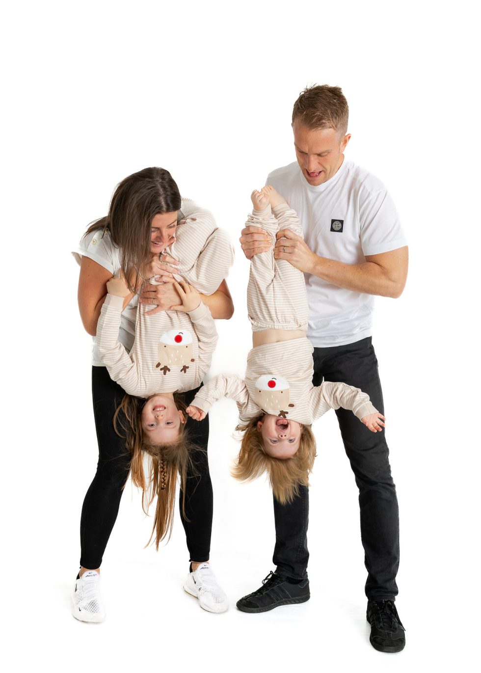 A smiling woman and man hold two laughing young children upside down. All wear light-coloured, matching pyjamas with reindeer faces. The background is plain white, creating a bright, cheerful family portrait full of playful energy.