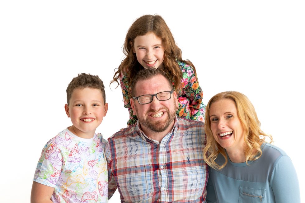 A joyful family of four poses against a white background. A man with glasses and a beard smiles widely in the centre, flanked by a woman with blonde hair, a young boy in a colourful shirt, and a young girl in a floral top who is standing behind them, all smiling brightly.