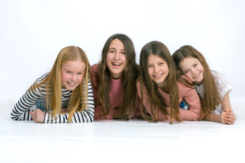 Four girls with long hair lie side by side on the floor, smiling at the camera. The background is white. The girl on the left wears a striped shirt, the next wears pink, and the other two wear pink and white tops, all appearing cheerful and relaxed.