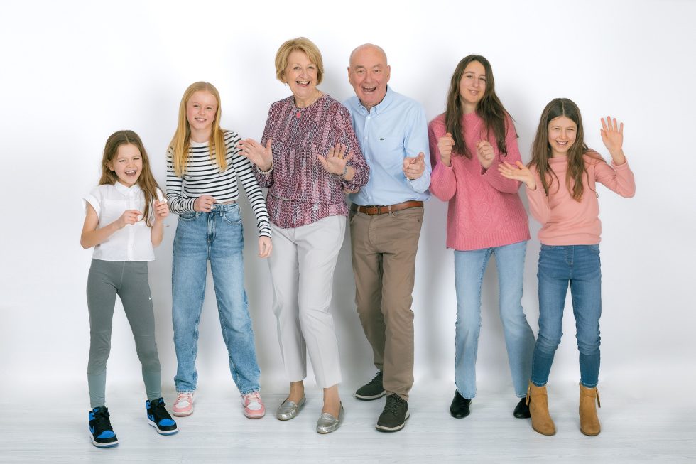 Six people—two older adults and four young girls—stand together in a row against a white background, smiling and posing energetically. The adults are in the centre, flanked by the children, all dressed in casual clothing and appearing joyful.