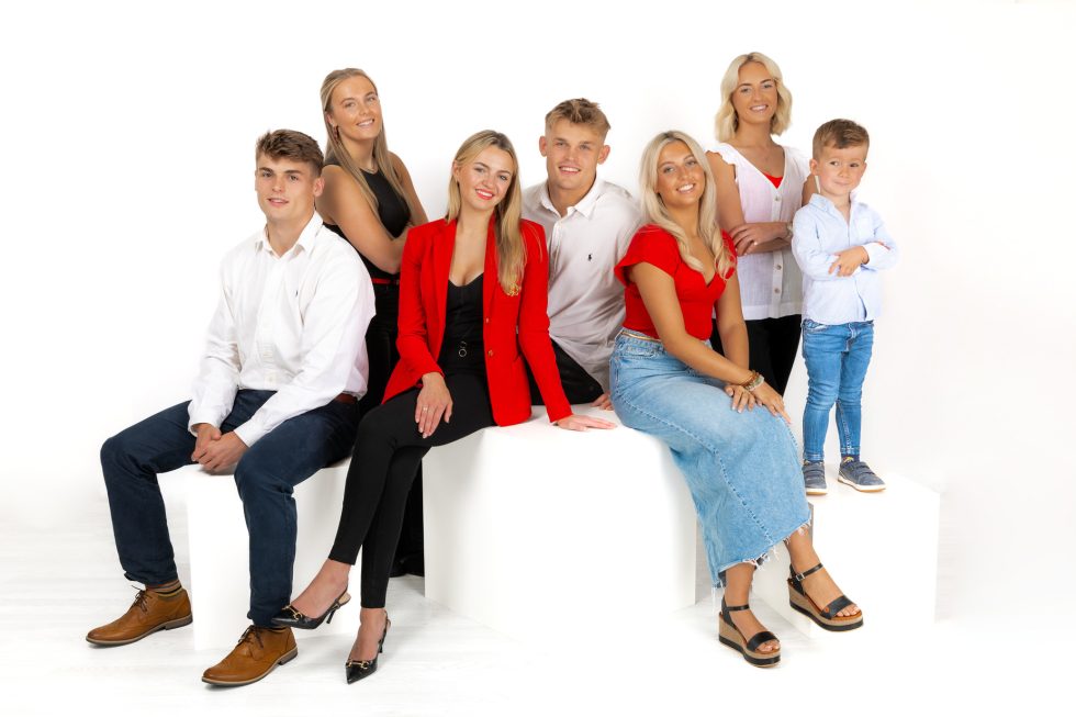 Seven people pose in a studio with a white background: three women in red, black, and white outfits, three men in white shirts and jeans, and a young boy in a blue shirt and jeans, all smiling and sitting or standing on white blocks.