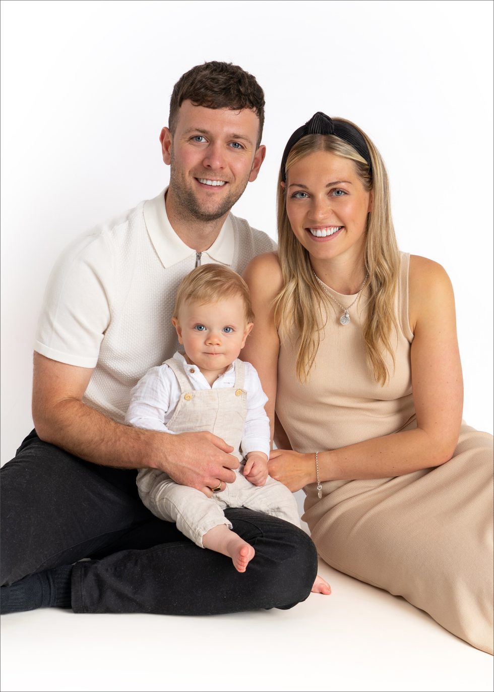 A smiling man and woman sit close together on the floor, both wearing light, neutral-coloured clothes. The man holds a baby in light dungarees on his lap. All three face the camera against a plain white background.