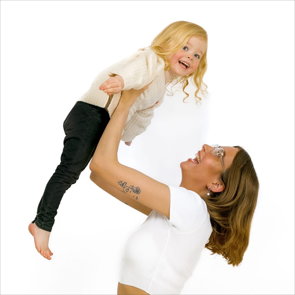 A smiling woman with a floral tattoo on her forearm lifts a happy barefoot blonde child in a cream jumper and black trousers against a white background. Both look joyful, making eye contact as the child stretches her arms out while being held.