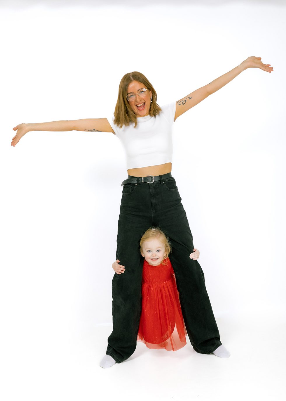 A smiling woman in a white crop top and black jeans stands with arms outstretched. A young girl in a red dress stands between her legs, holding onto them and smiling. Both are barefoot against a plain white background.